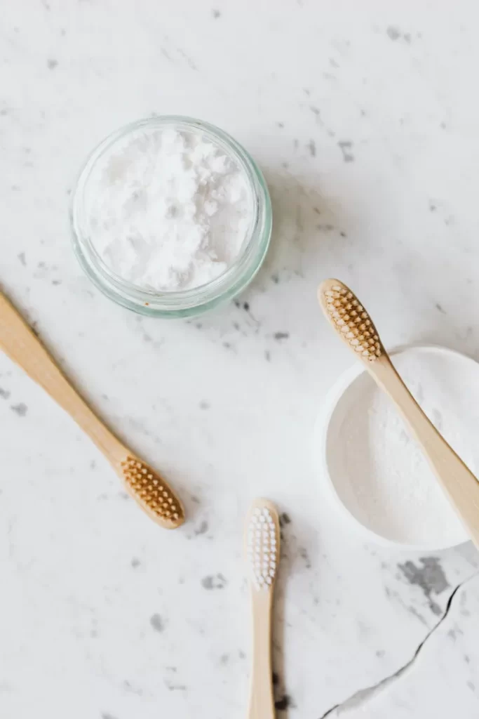 A jar of white powder, presumably baking soda, and a container with more powder are on a marble surface, alongside three toothbrushes with wooden handles