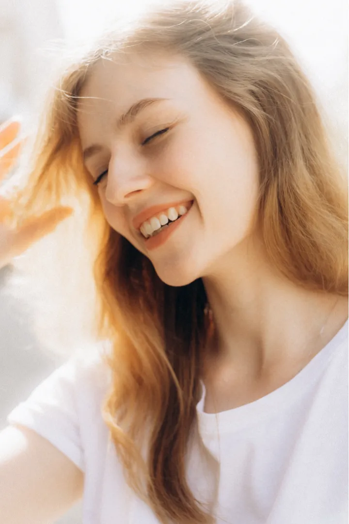 A woman with long, light brown hair smiles and squints her eyes while raising her hand, standing in bright sunlight