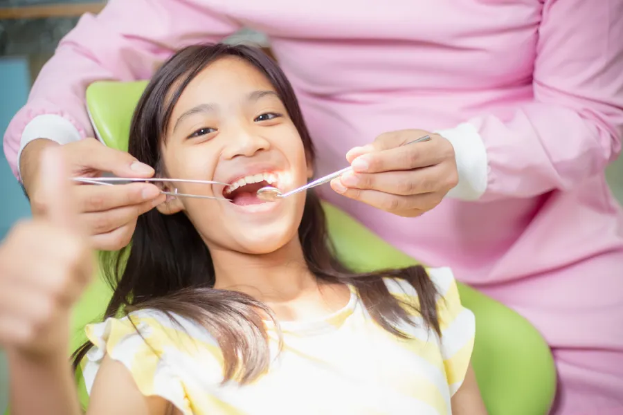 A young girl is sitting in a dental chair, smiling while a dentist checks her teeth